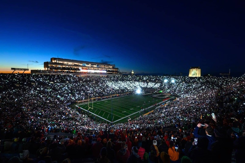 Notre Dame Stadium lit up at night by the fans phone flashlights. In the background, the Golden Dome and Touchdown Jesus glow against the dark sky.