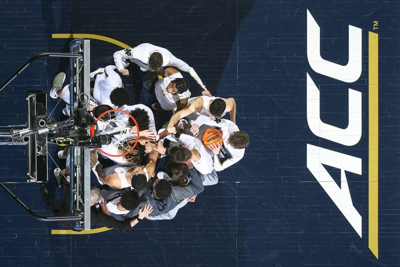 Notre Dame's Men's basketball team huddles underneath the basketball hoop. The ACC logo painted on the ground to the right of the team.