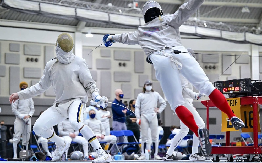 Two people fencing surrounded by other players or coaches.