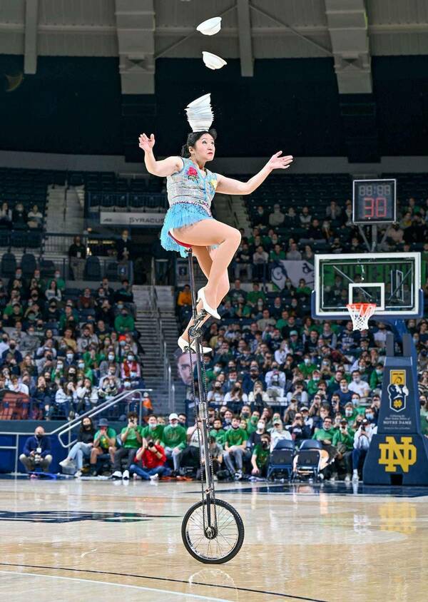 A woman rides on an extra tall unicycle balancing bowls on her head during a basketball halftime show.