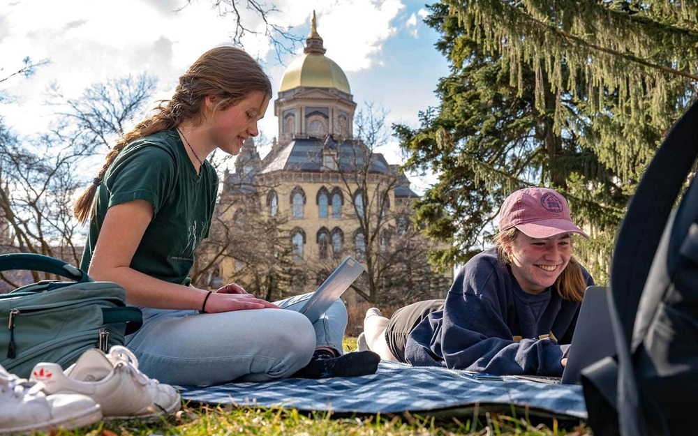 Two female students sit outside on blankets during a warm day. The golden dome in the background.