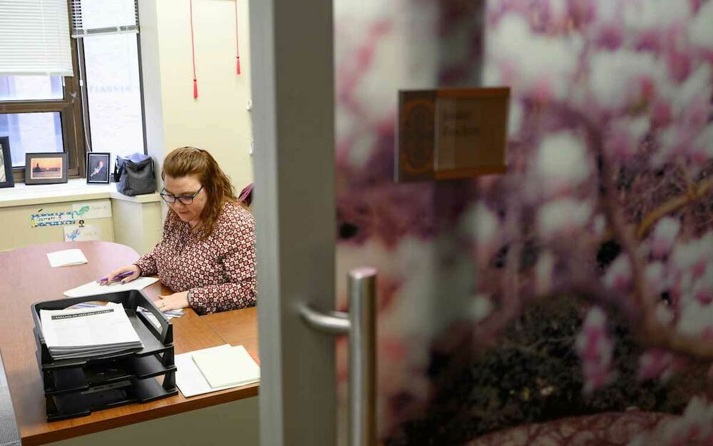 A woman, behind a cracked sliding door, sits at an office a desk and goes through paperwork.