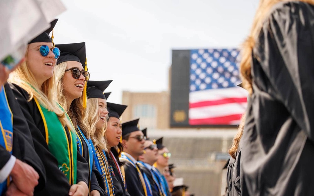 Graduates wearing caps and gowns standing looking right. In the background a stadium video board displays an American flag.