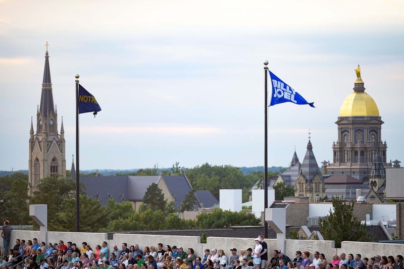 A Notre Dame flag flies next to a Billy Joel flag in the Notre Dame stadium. In the background the Main Building and Basilica can be seen.