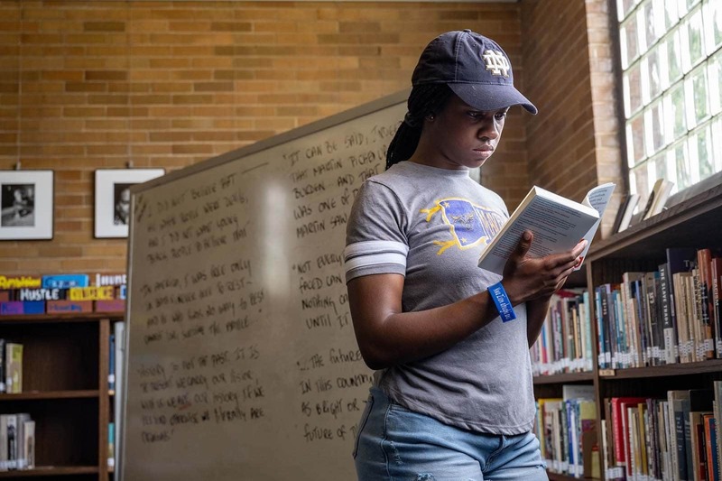 A Black woman wearing a Notre Dame hat stands in a room in front of a white board and reads a book.