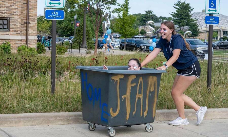 A female student pushes a large laundry cart with another female student peaking her head out from the top.