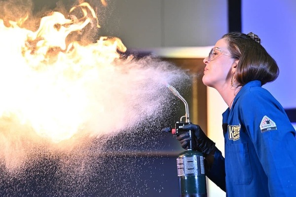 A young female in a blue lab coat and safety goggles stands to the right of the image and faces left. She  holds a gas canister and is seen spitting a flammable liquid into the air. A massive ball of fire fills the left side of the image.