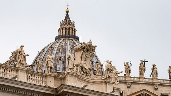 A close-up of the ornate roof of a large building with multiple statues, a dome, and a cross on top against a cloudy gray sky in Rome, Italy.