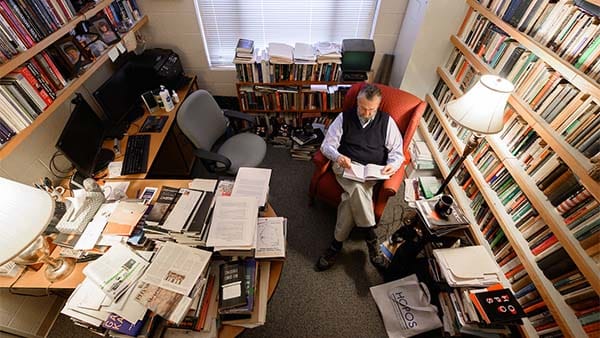 A professor sits reading a book in a cluttered office.