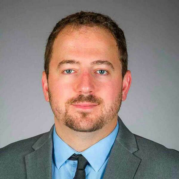Associate professor, Matt Webber wearing a gray suit, blue shirt, and black tie.