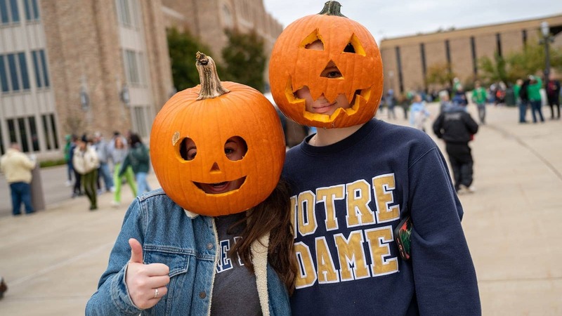 Two students in Notre Dame sweatshirts wear carved pumpkins on their heads.