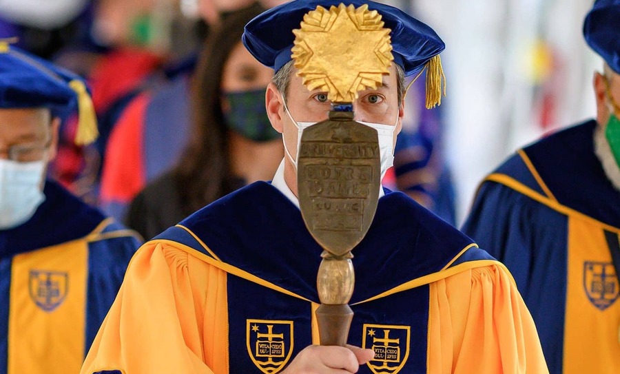 A masked man wearing a cap, hood, and gown holds a mace in front of his face.