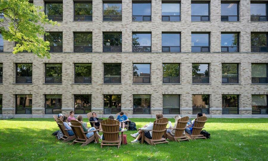 A class forms a circle sitting in Adirondack chairs in front of a windowed building.