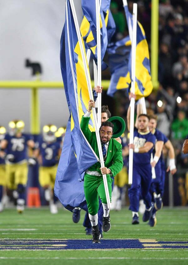 A Leprechaun mascot runs onto the stadium field holding an ND flag, followed by cheerleaders and the football team.