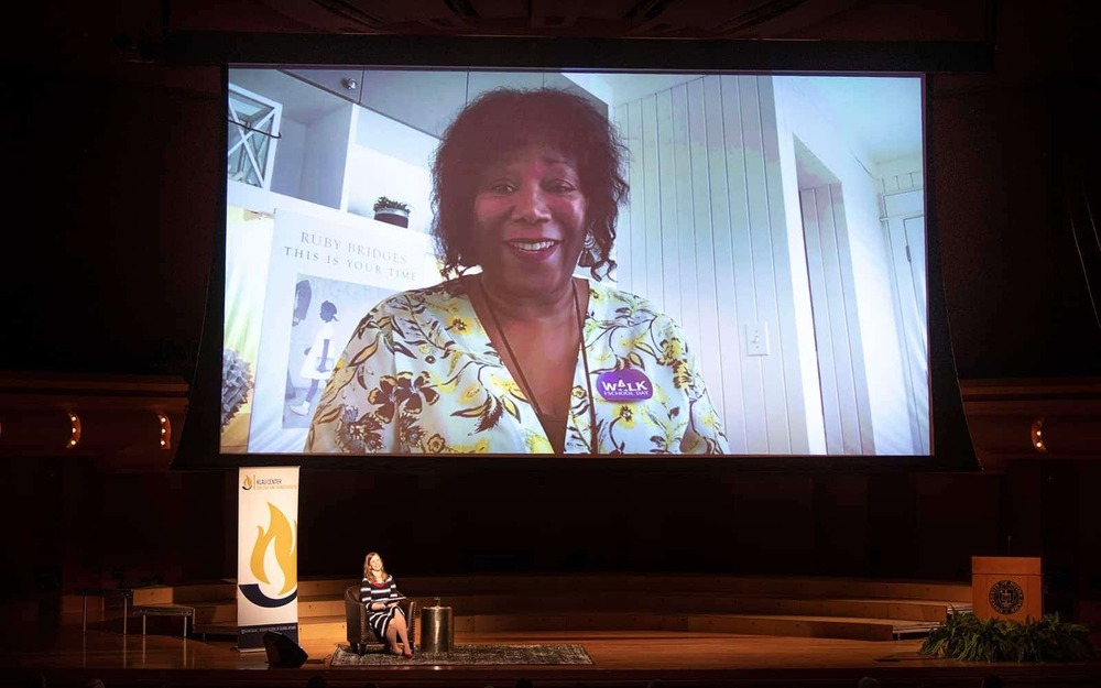 A woman sits in a chair next to a Klau Center retractable banner. Behind her is a large projected screen with civil rights icon Ruby Bridges on video.