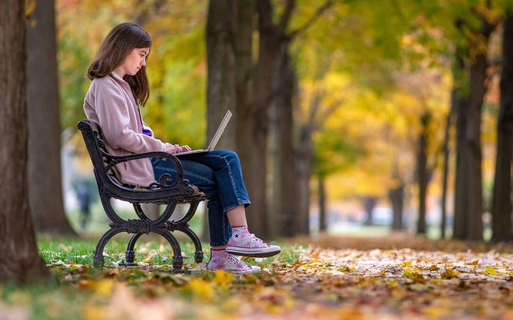 A student sits on a park bench next to a sidewalk covered in fall leaves.