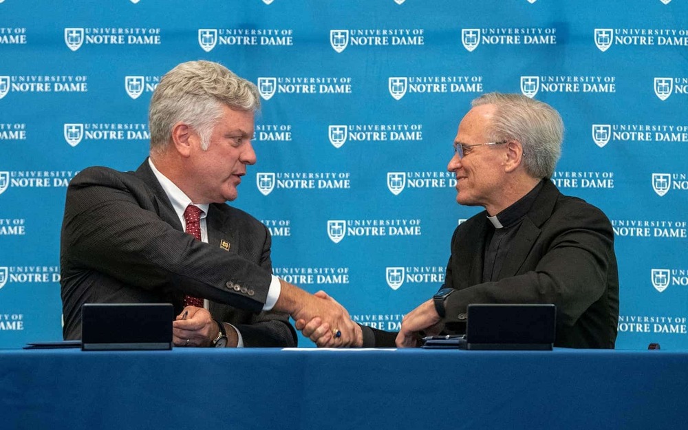 Steven H. Walker and Rev. John I. Jenkins, C.S.C. sit behind a University of Notre Dame logo backdrop and shake hands.