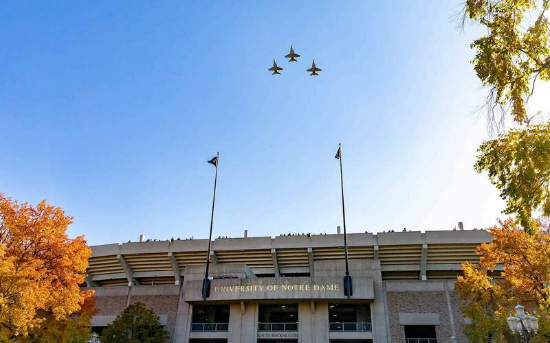 Three U.S. Marine Corps F/A-18 Hornet airplanes fly over Notre Dame Stadium. The sky is clear and blue and the trees off to the side of the photo are yellow.