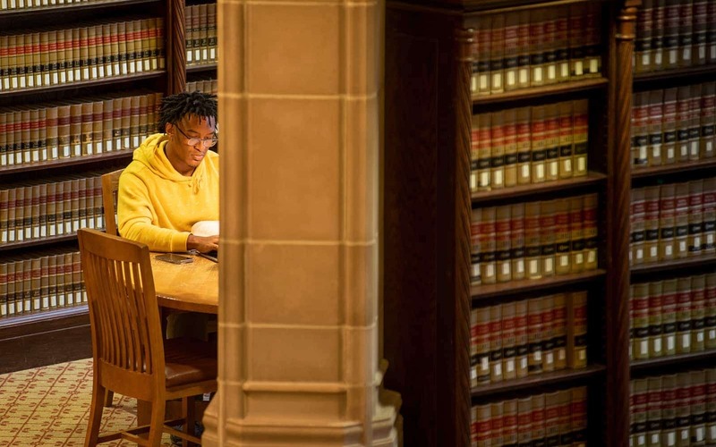 A student studies at a desk between book shelves in a library.