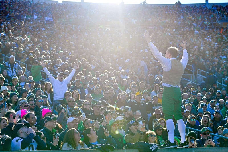 The Notre Dame Leprechaun stands in front of Notre Dame fans sitting in the stadium and urges everyone to cheer and get excited.