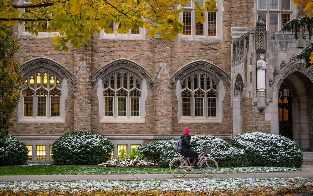 A student rides a bike past a campus building. Light snow covers the nearby grass and bushes.