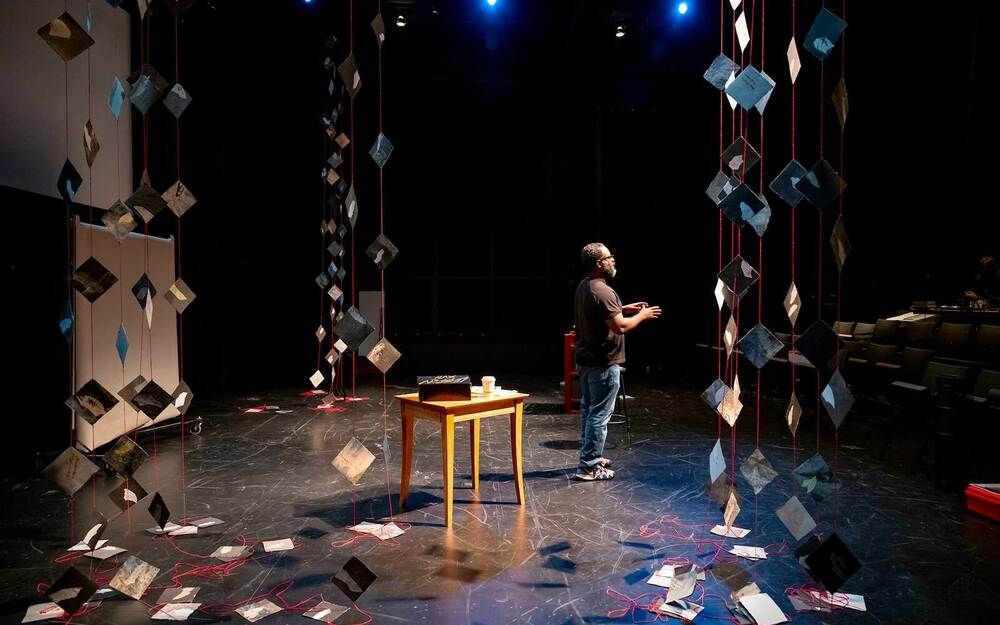 A man stands on a dim stage. Surrounding him are printed papers held by red string from the ceiling and draping down to the floor.