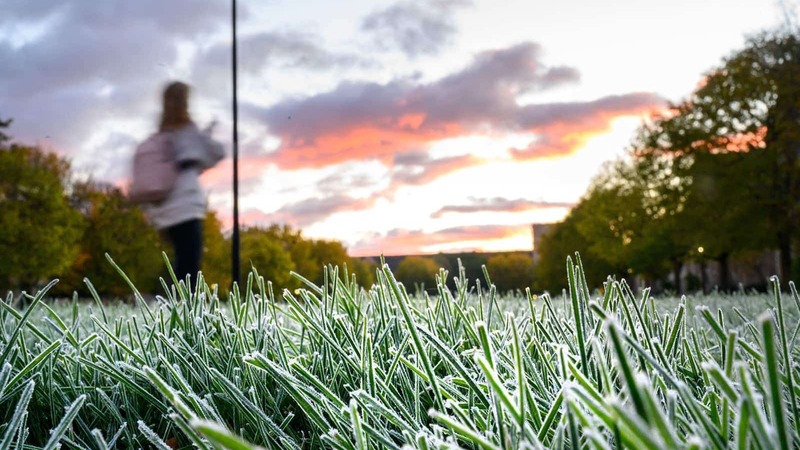 Grass covered in frost as the sun rises and one student walks by in the background.