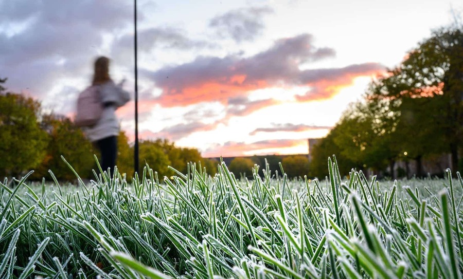 Grass covered in frost as the sun rises and one student walks by in the background.