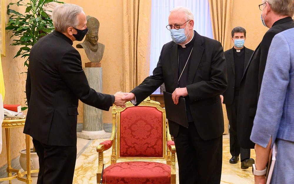 Two priests wearing masks shake hands, other people stand around them.