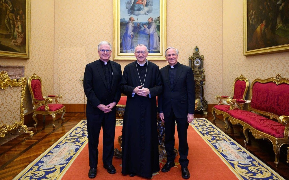 Two priests and cardinal priest stand in a room and post for a photo. There are ornate furniture, rugs, and artwork around them.