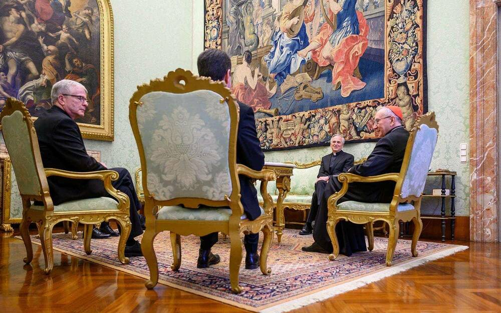Four people sit in ornate and antique chairs. The walls are lavishly decorated with artwork.