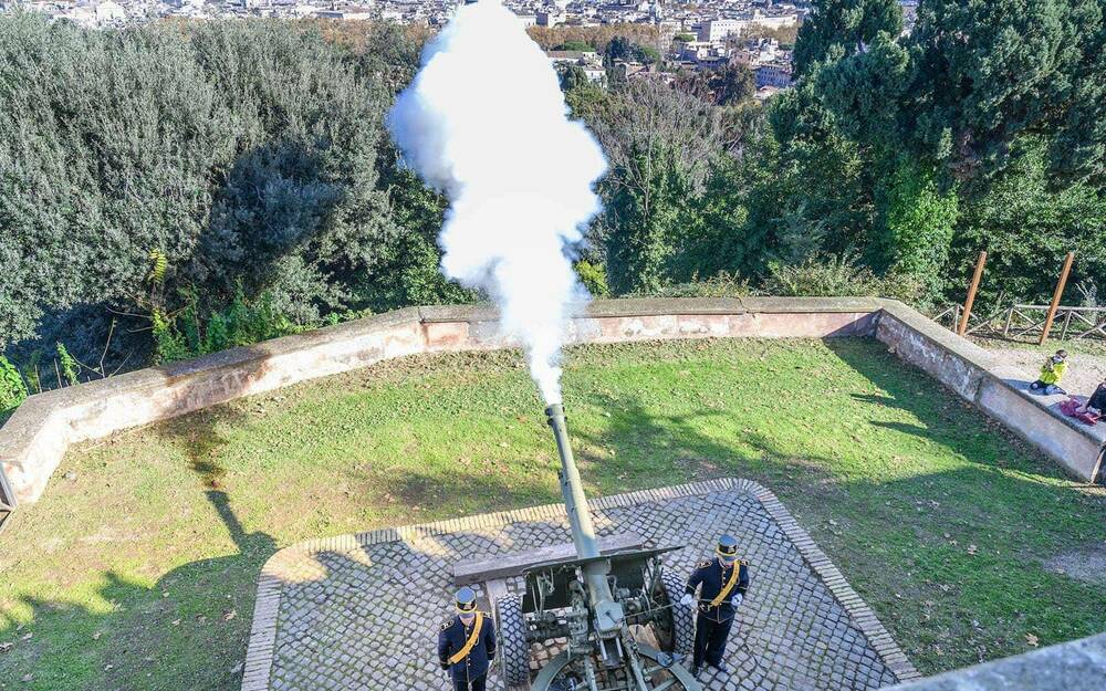 Smoke billows out of a canon outside in a yard. Two guards stand on each side of the canon.