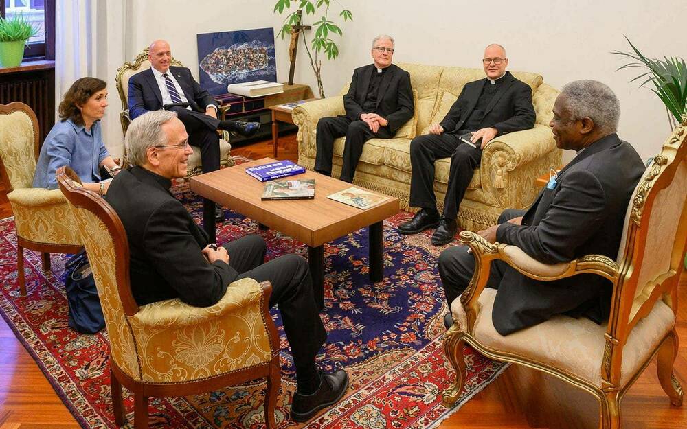 Six people, including four priests, meet sitting on ornate and antique chairs.