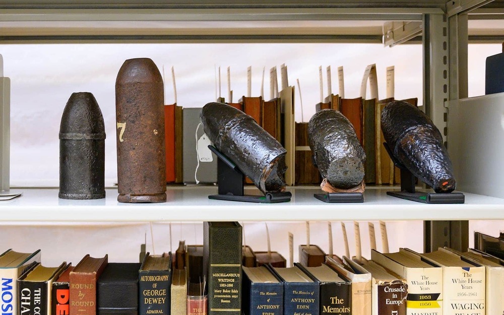 Artillery shells on a top shelf of a bookshelf, a collection of books below it.