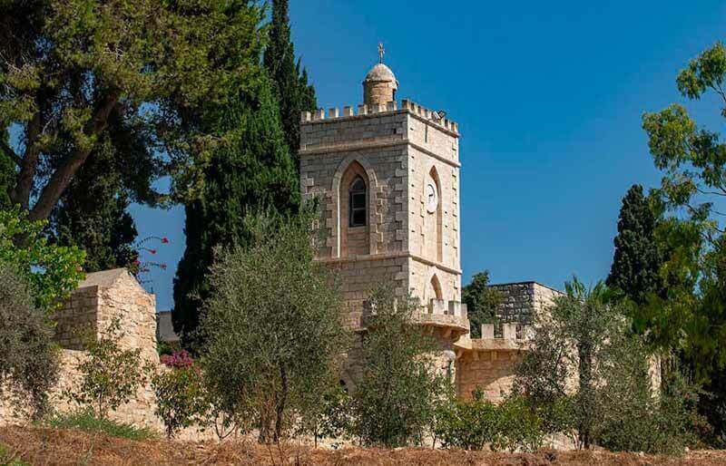 A stone tower with a small cross on top stands among a cluster of trees against a clear blue sky.