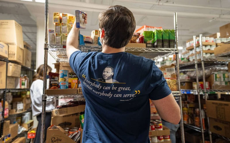 A student reaches for a boxed meal on a top shelf. The back of their shirt states a Martin Luther King Jr quote: 'Everybody can be great, because everybody can serve'.