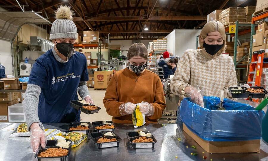 Three masked students stand side by side assembling meals in divided food trays.