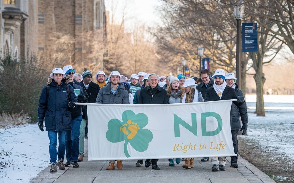 A group of people wearing white bucket hats march through campus holding a larger banner that says 'ND Right to Life'.