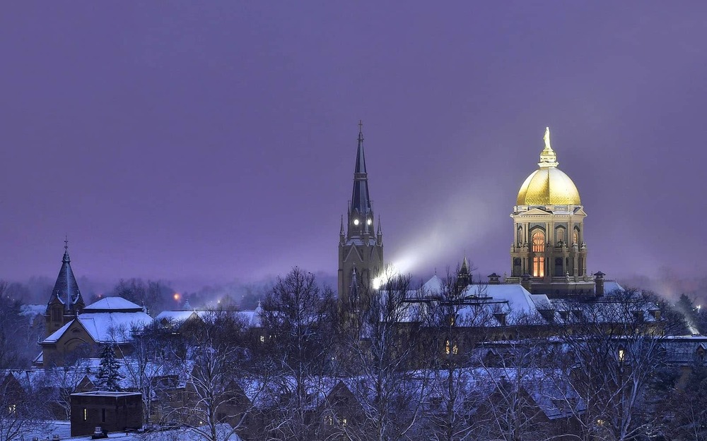 Lights shine brightly onto Notre Dame's Golden Dome during a snowy dusk evening.
