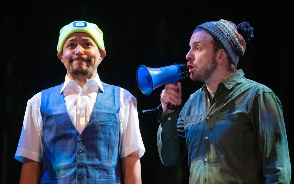 Two actors on a stage, one talks into a megaphone towards the other who makes a concerning face.