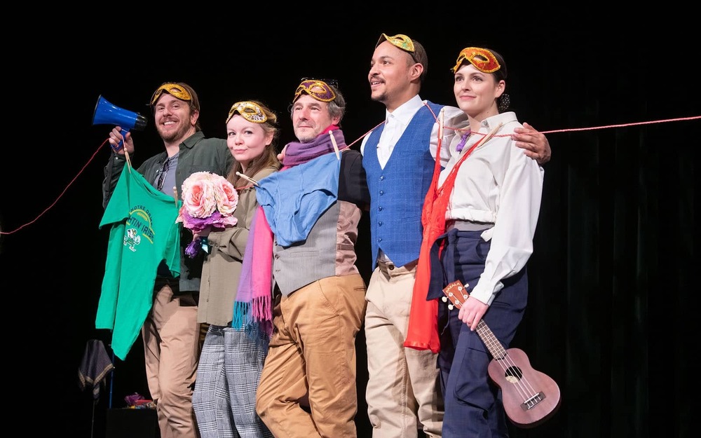 A group of people on stage tied up in a clothes hanger smile and smirk. There are pieces of clothing hanging on the line. One actor holds a ukulele and another hold a megaphone.