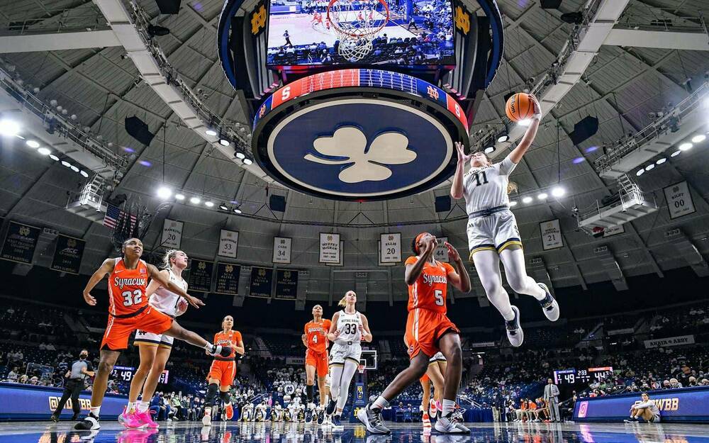 A Notre Dame women's basketball player jumps in the air for the ball. Others surround her on the floor.
