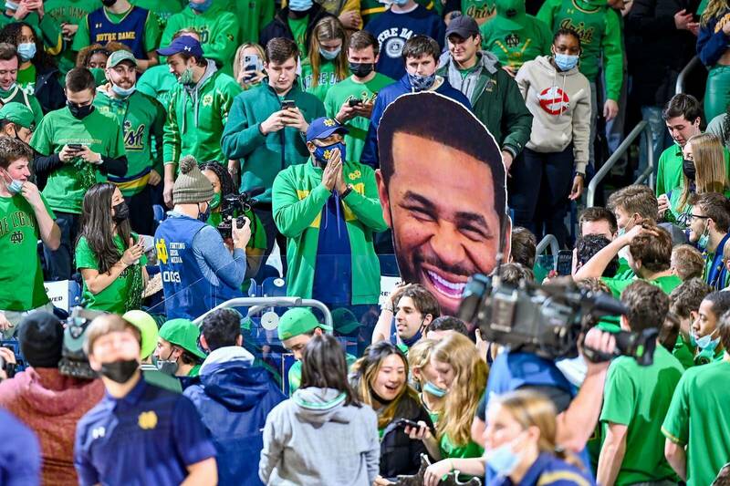 Former Notre Dame football player and NFL Hall of Fame enshrinee Jerome Bettis is surrounded by students and fans in the stands during a Men's basketball game. A student holds onto a large fat head of Jerome next to him.