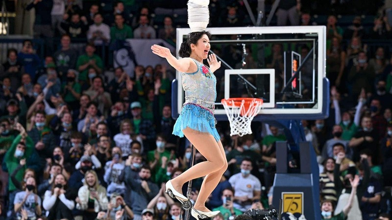 A woman rides on an extra tall unicycle balancing bowls on her head during a basketball halftime show.