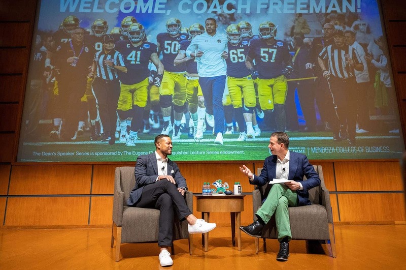 Two people sit in chairs on stage and have a conversation. An image of the football team and coach running out of a tunnel projected behind them.