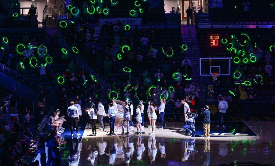 A basketball court in darkness. A light shines on the team and neon green circle lights shine into the crowd.