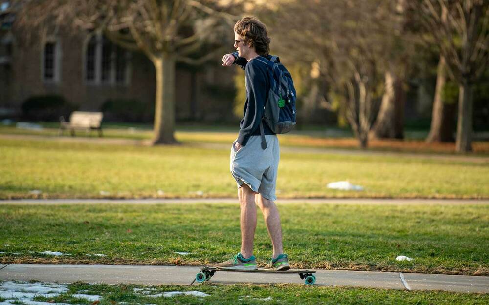 A student skateboards on a sidewalk while talking on his smart watch.