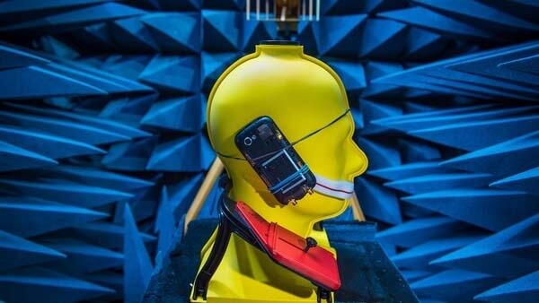A yellow mannequin head with a mobile phone attached to its side is positioned in an anechoic chamber with blue foam spikes on the walls.