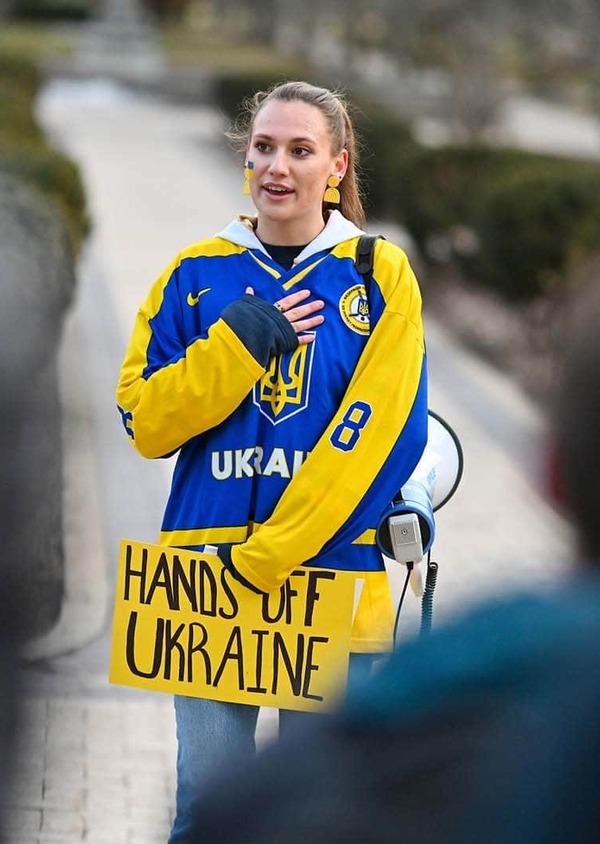 A student wearing a Ukrainian jacket and holding a sign that says 'Hands off Ukraine' speaks to other students on campus.