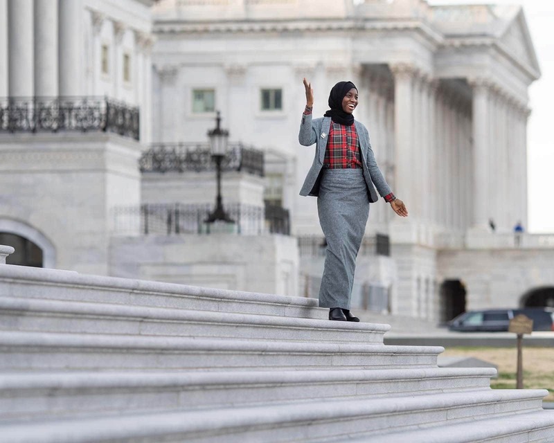 A woman wearing a Hijab stands on the steps of the US Capital Building and waves.
